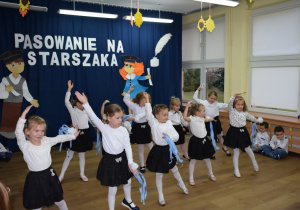 Na tle dekoracji dzieci stoją w półkolu, na pierwszym planie dziesięć dziewczynek tańczy układ taneczny z niebieskimi szarfami